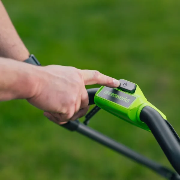 Fingur á ræsihnappi á handfangi Greenworks 36 cm sláttuvélar.